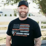 Smiling bearded man in Grand View Roofing shirt and baseball cap outdoors