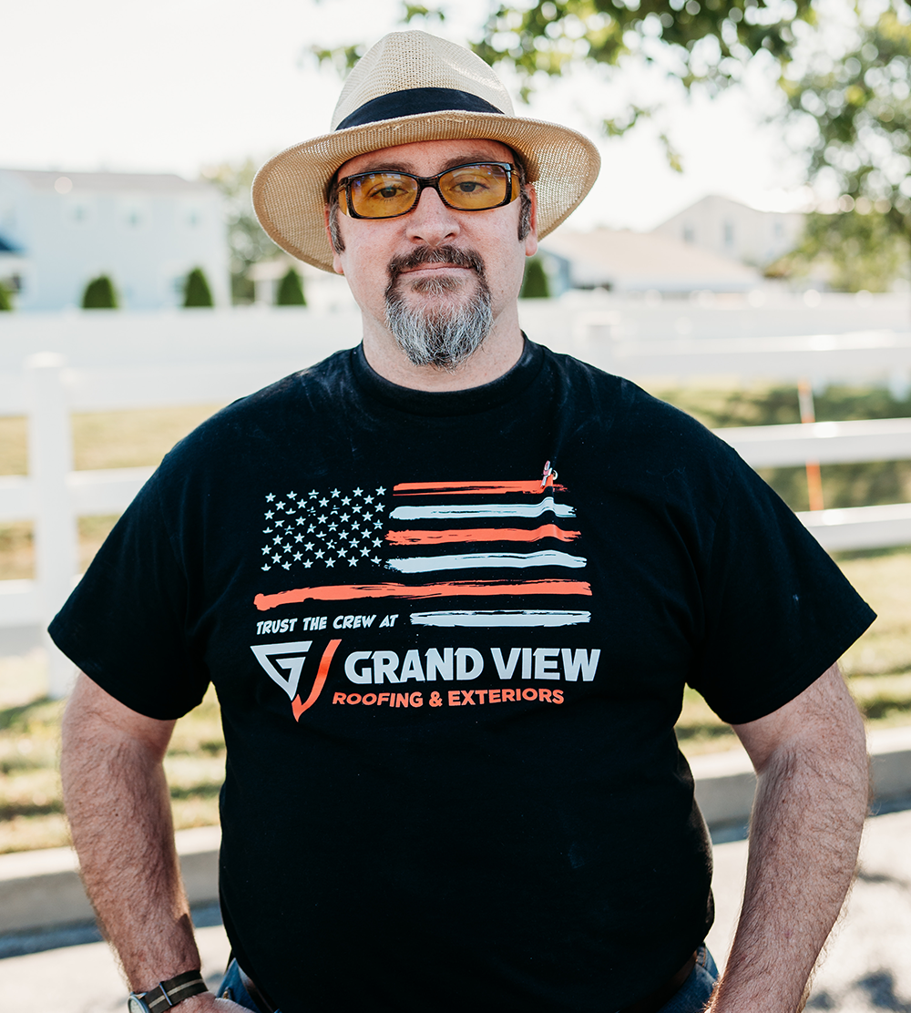 Man in hat and glasses wearing Grand View Roofing & Exteriors t-shirt outdoors
