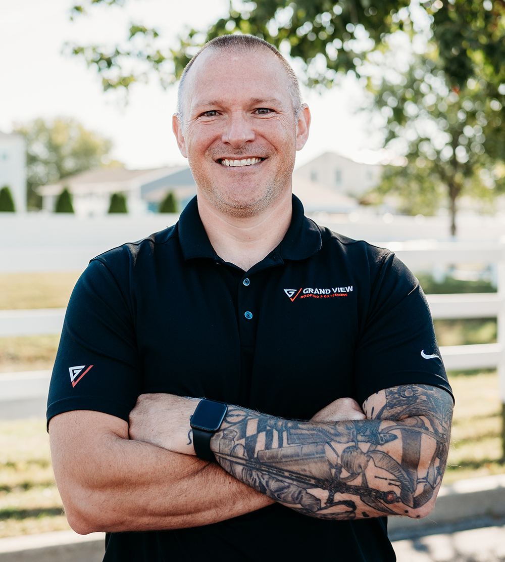 Smiling man with tattoos in navy polo shirt standing outdoors with arms crossed